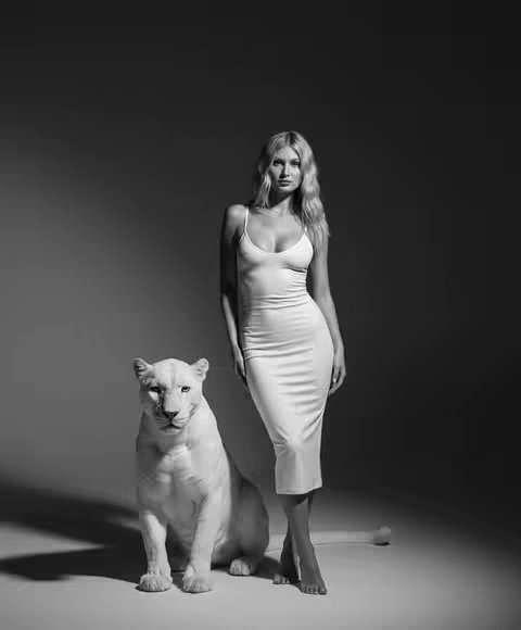 A black and white photograph depicts a woman in a white dress standing near a white lion. The woman has long blonde hair and is looking towards the camera. The lion is seated beside her, also in a seated pose. The background is a muted gray. The lighting highlights the textures of the dress and the lion's fur. The composition is centered, using the subjects to fill the frame and the woman looks elegant giving off a sense of sophistication.