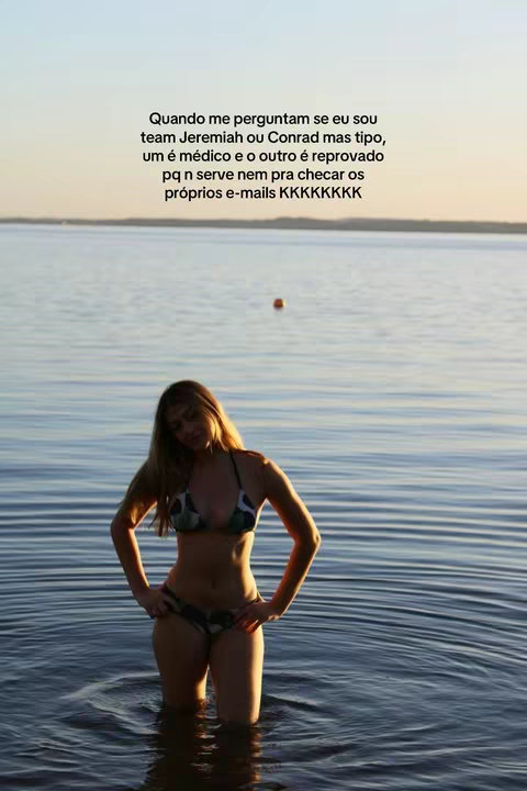 A woman stands in waist-deep water, facing the viewer with hands on her hips. She wears a bikini and the background shows a calm lake and sky.