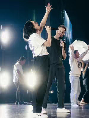 A live performance shot featuring two individuals on stage. The focus is on a woman with short dark hair, wearing a white t-shirt and black pants, singing with her arm raised and a microphone in her hand. Beside her is a man wearing a black shirt and pants, also holding a microphone. The lighting is stage lighting, with other people with white clothing in the background and stage lights creating a blue-green hue.
