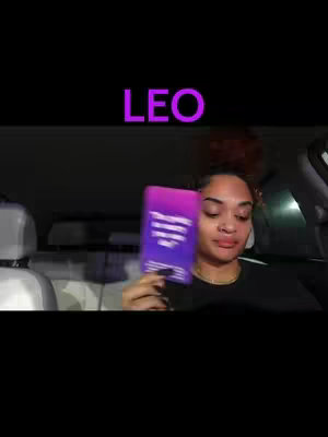 The image shows a person in a car holding up a tarot card. The word "LEO" is written in large, purple, stylized letters above the person. The person is a woman with dark hair and red highlights, wearing a black shirt, with her eyes focused directly at the camera. The background shows the interior of a car, with a neutral color palette dominated by blacks and beiges.