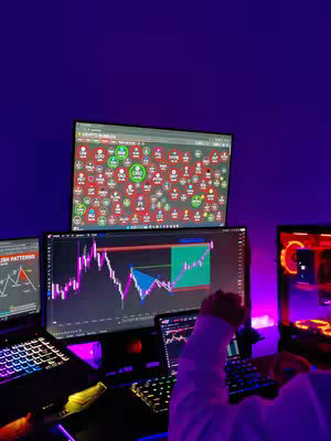 The image shows a dimly lit room with multiple computer screens showcasing trading charts and data. In the foreground, a person's hand is visible, interacting with a keyboard, suggesting active trading or analysis. The screens display various data visualizations and trade-related information, indicative of a trading or financial analysis setup. The lighting is moody, with purple hues dominating the scene, which creates an atmosphere of focus and intensity. The multiple screens and computer equipment suggest a professional or dedicated environment for trading.