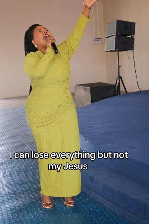 The image features a woman in a bright green dress, standing in what appears to be a stage setting. She is holding a microphone and has one arm raised, with an expression of joy or praise on her face. The background includes sound equipment and a blue stage, and across the bottom, white text provides a message.