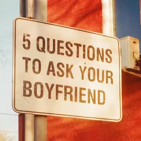 A rectangular, weathered road sign with the words '5 QUESTIONS TO ASK YOUR BOYFRIEND' in a bold, shadowed font. The sign is mounted on a silver pole in front of a textured, red wall. The sign is slightly at an angle, and in the background, there's a glimpse of a blue sky and an electrical box.