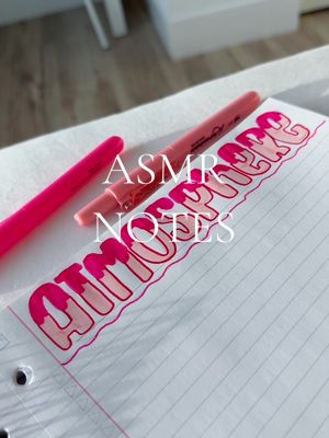 The image presents a close-up view of a notebook page featuring the handwritten title "ATMOSPHERE NOTES" in a bubble letter style. The letters are primarily pink with a darker pink outline, and there are two pink pens placed to the side of the title. The image is well-lit, with a clean and simple composition.