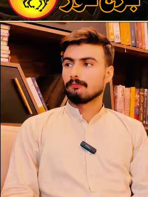A man with a beard and short hair, wearing a light-colored shirt, is the central focus. He looks slightly off-camera. Behind him is a cluttered bookshelf and a small microphone clipped to his shirt.