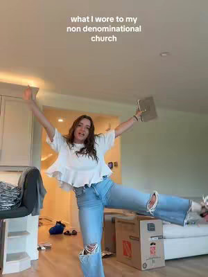 A woman with long brown hair is in a light domestic space with her arms outstretched. She's wearing a white top, ripped jeans and holding a book. Cardboard boxes and other objects are in the background.