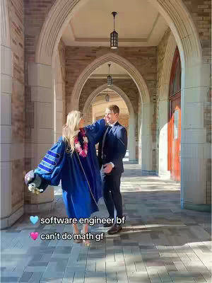 A couple stands in an arched hallway. The woman is wearing a graduation gown and holding a bouquet of flowers, while the man in a suit is smiling and looking at her.
