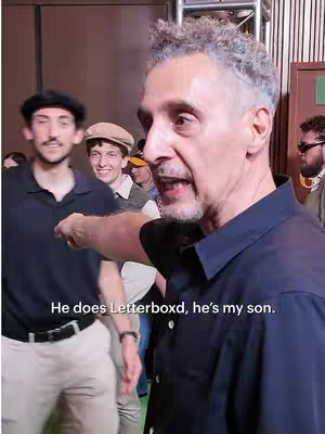 A medium shot features three men in a setting that appears to be indoors, possibly a behind-the-scenes look. The man on the right has graying curly hair, a dark short-sleeved shirt, and is pointing with his right arm, seemingly addressing someone not visible. To his left are two younger men, one in a black shirt and the other in a tweed cap. All are in focus with a blurred background.