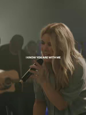 A vertical video cover shows a woman singing into a microphone. Overlayed at the top is the text 'I KNOW YOU ARE WITH ME'. The background is dim and appears to be a stage or studio setting, with several indistinct figures visible.