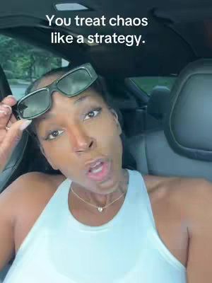 A woman with sunglasses sitting in a car, speaking to the camera. The background is blurred, suggesting a parked vehicle.
