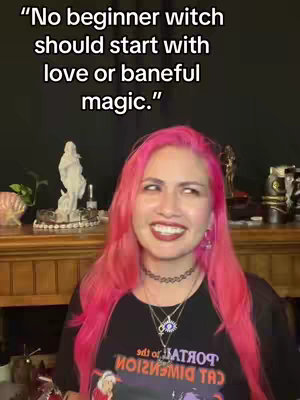 A woman with pink hair is smiling, looking slightly to the side. Text in a black font, "No beginner witch should start with love or baneful magic." is overlayed at the top of the image. The background includes wooden furniture and a statue.