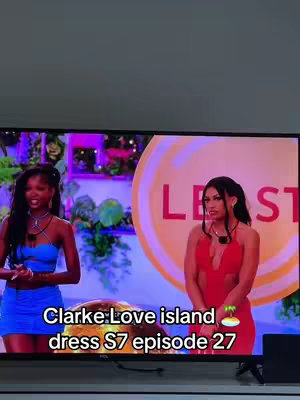 A video cover showing two women on a screen. Text overlay at the bottom identifies a specific dress. The background shows a blurred out setting with some colorful elements, making the women the focal point.