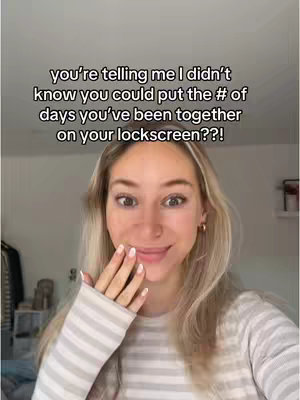 A young woman with blonde hair and a light-colored striped shirt is centered in the frame, looking at the camera with wide eyes and a slight smile while holding her hand up near her face. Overlayed text appears in the top half of the frame and says "you're telling me I didn't know you could put the # of days you've been together on your lockscreen??!?"