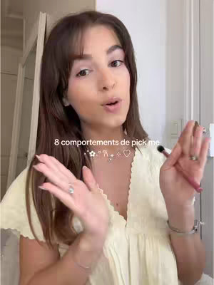 A young woman with light makeup and long brown hair is centered in the frame, facing the camera. She wears a lightly colored, ruffled dress. Above her, there is a text overlay featuring a title and decorative symbols.