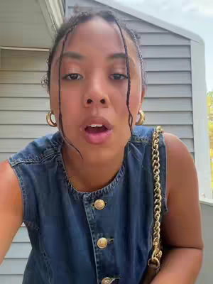 A close-up shot of a young woman with dark hair, braided into small sections, and wearing gold hoop earrings. She appears to be speaking, with her mouth open. She's wearing a denim dress with gold buttons. The background shows a section of a gray wooden exterior wall.
