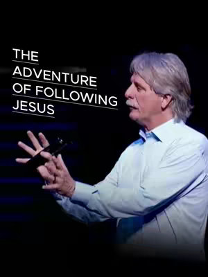 A man with gray hair and a light blue shirt, holds a microphone and gestures towards the audience. The text overlay says: 'THE ADVENTURE OF FOLLOWING JESUS'. The background is dark.