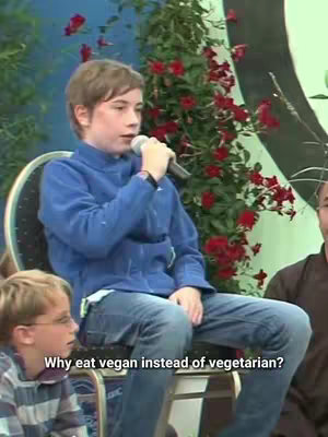A young boy speaks into a microphone while seated. The background features a vibrant display of red flowers and greenery.