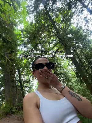 A selfie taken in a forest setting with a woman in the foreground. She is wearing sunglasses and a white top, hand covering her mouth. Background is blurred trees and foliage.