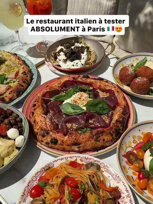An overhead shot of a white table features numerous Italian dishes: a large pizza in the center with a red rim and toppings, a pan of what appears to be pasta with truffle, a bowl of olives, and a plate of what appears to be pasta with clams.
In the background, a person can be seen drinking from a glass of orange liquid which suggests an Aperol spritz is present.