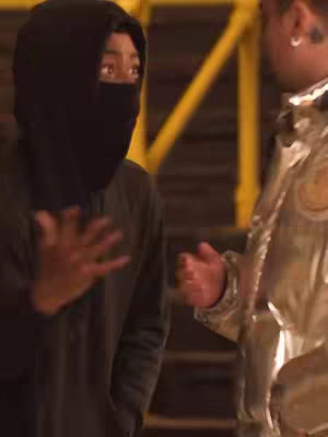 The image shows two young men interacting in what seems to be an urban environment. The man on the right is wearing a metallic jacket and has a serious expression. The man on the left is wearing a black hoodie and face covering. He extends his hand with an open palm, suggesting a gesture of communication or questioning. The background shows a glimpse of yellow support beams, suggesting a structure, possibly for filming.
