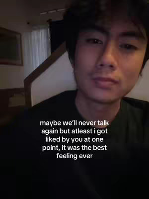 A selfie-style video cover image features a young man in a dimly lit room. The man, with dark hair and a slightly soft expression, is positioned in the center of the frame. The background includes parts of a room, a wall and window, the lighting seems to be indoor. Text overlay in white font appears toward the bottom, expressing a melancholic sentiment.