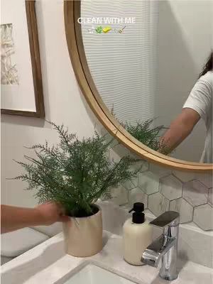 A person is setting down a plant in a bathroom. The bathroom has a round mirror, white walls, and a white sink. The person's reflection is visible in the mirror. The title 'CLEAN WITH ME' is overlaid at the top of the image. The overall aesthetic is clean and modern.