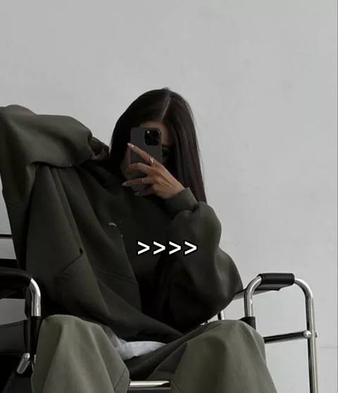 The cover image features a woman sitting in a wheelchair. She wears a dark green hoodie and dark pants, obscuring her face with a phone, with only part of her face and her hand holding the phone visible. The background is a plain white wall. Overlaid on the image is the text '>>>>' .