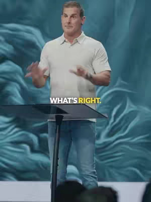 A man in a light-colored shirt and blue jeans stands behind a podium, gesturing with his hands as if speaking or presenting. The background has a blurred, textured teal appearance. The image is well-lit, with the man being the central focus.