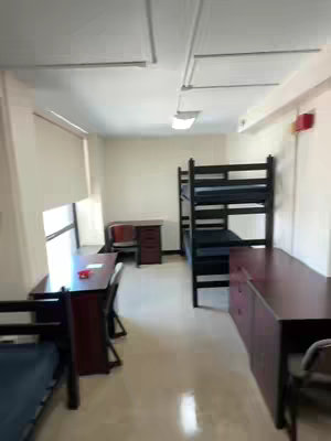 The image shows a dorm room, with bunk beds on the right side of the frame, desks, and a window with blinds. The room is lit by fluorescent lights, with a dark red desk and furniture. The color palette is generally light.