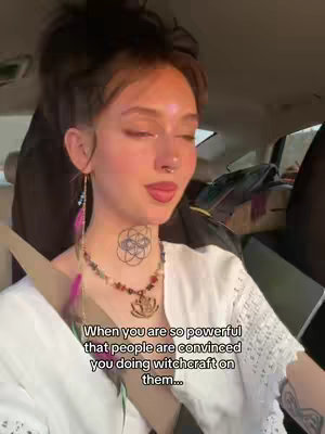 A young woman with dark hair in a messy bun is seated in a car. She has light skin, a nose ring, and visible makeup. She is wearing a white top and a colorful necklace with a lotus pendant. Text overlayed on the image reads, 'When you are so powerful that people are convinced you doing witchcraft on them...'