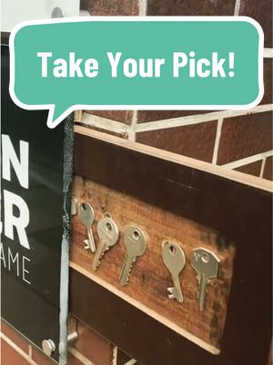 The image shows a close-up of a wooden surface with keys and a speech bubble. The surface is made of dark brown wooden panels and is decorated with five silver keys, each hanging by its hole. A teal speech bubble at the top left says \"Take Your Pick!\" in white text. Behind the wooden panel is a texture of bricks.