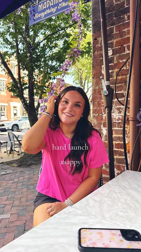 A young woman with long dark hair is smiling at the camera while sitting outside a restaurant. She’s wearing a pink t-shirt and has bracelets. The background shows trees, a sign, and a brick wall.