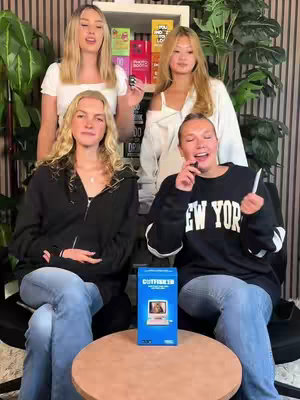 The image features a group of four young women, seemingly in a studio setting. They vary in appearance, with diverse hairstyles and clothing. One woman holds a product box. The setup includes a background with vertical lines and shelves full of products.
