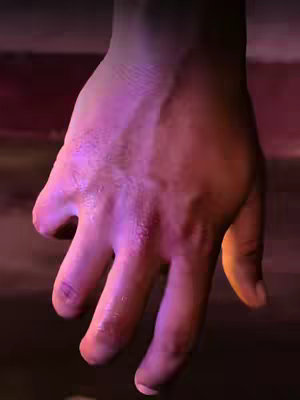 A close-up shot of a hand, palm facing up. The hand is partially lit with a gradient of colors, with the upper part of the hand in a purplish hue and transitioning to orange near the wrist, creating a dramatic effect. The background is dark, which emphasizes the hand as the main focus. The lighting gives the image a moody and emotional feel.