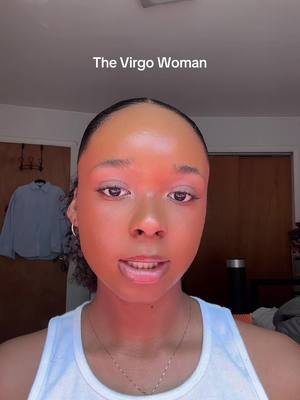 A close-up shot of a woman's face dominates the frame, with warm lighting setting a slightly reddish tone across her skin. The text overlay reads "The Virgo Woman" in a straightforward sans-serif font. The woman's eyes are open, and she appears to be speaking, with her lips slightly parted. She wears a simple white tank top and a delicate gold necklace. The blurred background shows what appears to be a bedroom or living space with a door and some clothing items, a closet door or a similar structure is also visible.
