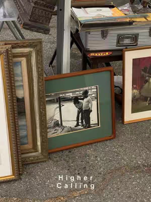 The image features a collection of framed pictures displayed on a rough, gray surface, possibly a concrete floor. The composition focuses on the arrangement of the frames, with the corner of a few paintings and a framed picture depicting people. At the bottom, text saying “Higher Calling” is displayed.