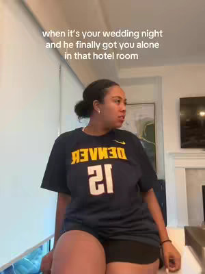 A woman wearing a Denver Nuggets jersey stands in what appears to be a hotel room, looking off to the side with a concerned or surprised expression. The room is brightly lit, with white walls and a visible window. The text overlay suggests a humorous scenario related to a wedding night.