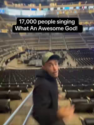 A man in a black hoodie and green cap is standing in an arena, looking at the camera. A black text box in the top left of the image reads: '17,000 people singing What An Awesome God!' The background shows stadium seating.