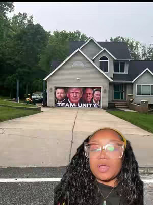 A woman with glasses and long curly hair is standing in front of a house with a banner that displays “TEAM UNITY” with pictures of several men on a banner that covers the garage door. The woman is in the foreground with an open mouth.