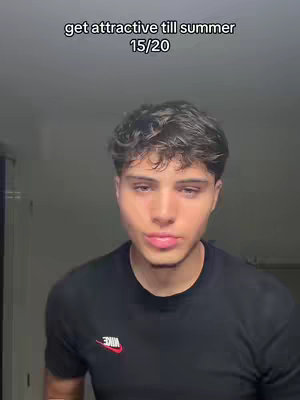 A young man with dark, wavy hair looks directly at the camera in a selfie-style shot. He's wearing a black t-shirt with a white and red Nike logo. The background is a blurry, solid light color. Text at the top reads 'get attractive till summer 15/20'.