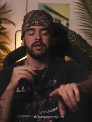 A man with closed eyes wearing a camouflage bandana is seated in a chair, with a dark background. He has a beard and a necklace, and he is wearing a black shirt. Palms and greenery are in the background.