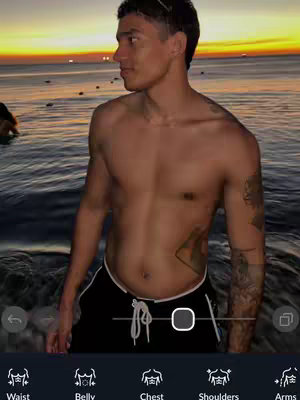 A shirtless man stands with his side to the camera at the edge of a beach or shore during sunset. The sky is orange, and the water has some reflection of the sunset. There are app features at the bottom of the image.