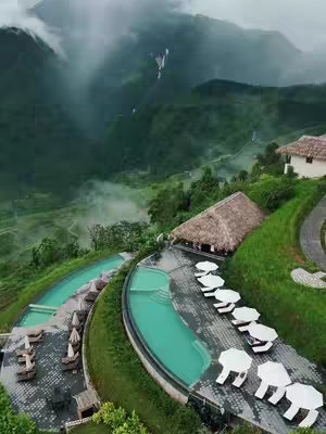 An aerial view showcases a lush green landscape with a resort-style area. The scene features terraced greenery that slopes into the valley. A sparkling pool with curved edges catches the eye alongside white lounge chairs and umbrellas. Houses with thatched roofs are nestled in the terrain and the whole scene is bathed in a soft, natural light with distant mountain views.