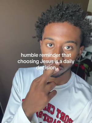 A medium shot featuring a young Black man looking directly at the camera. He has curly black hair and is wearing a white t-shirt. The text overlay is centered, and he has his index finger pointing at his lips. The background is blurred, with a beige neutral color.