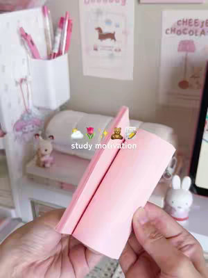 The image shows a pink notepad held in hands. In the background, a decorated desk is visible with study supplies. The aesthetic is soft and pastel-colored.