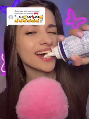 A young woman in a close-up shot is holding a whipped cream dispenser and applying the cream to her mouth. She is smiling and looking to her right with a commentary bubble overlay on the top left which contains text in Russian and different emojis (spoon, pot, bar). Behind the girl is a pink microphone and a pink butterfly neon sign. The lighting is diffused and soft, giving the image a warm feel.
