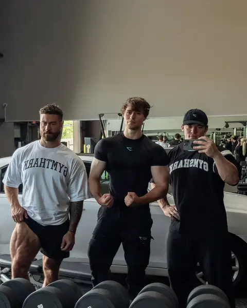 Three muscular men posing in a gym setting. The man on the left is wearing a white t-shirt and black shorts, showcasing his leg muscles. The man in the middle is wearing a black shirt and pants. The man on the right is taking a selfie.  The background includes gym equipment, a white car, and a mirror.