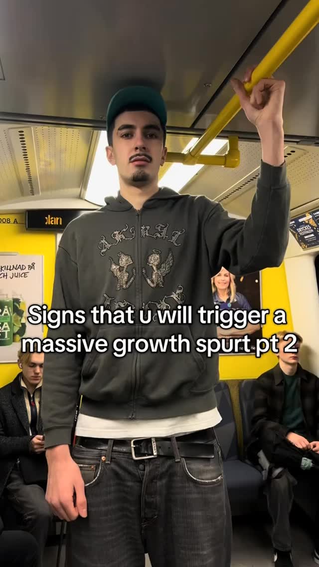 A man in a dark green hoodie stands on a moving train, holding onto a yellow pole. Text overlays read “Signs that u will trigger a massive growth spurt pt 2.” The background shows other passengers and advertisements. The color tone is cool, with a slightly desaturated appearance.