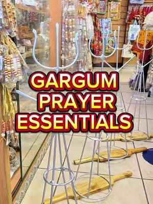 A photograph captures a retail setting showcasing religious items. The foreground features several stands with metal frames, each topped with a wooden item. A text overlay reads 'GARGUM PRAYER ESSENTIALS' in bold letters. In the background, shelves display various religious artifacts and decorations.