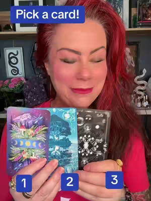 A person with red hair holding three tarot cards, with the text "Pick a card!" in a blue banner at the top. The person is smiling, and numbers are placed on the cards.